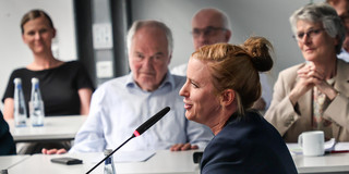 Side profile of Prof. Petra Wiederkehr at table with microphone, members of the University Election Assembly are seated in the background.
