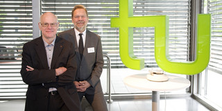 Zwei Männer lehnen an einem Tisch. Rechts neben ihnen auf dem Tisch steht ein großes grünes TU-Logo.