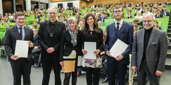 Till-Moritz-Karbach_Dissertationspreis Vier Männer und zwei Frauen stehen neben einander. Zwei Männer und eine Frau halten ein Dokument in der Hand. Im Hintergrund sitzen Menschen im Hörsaal.