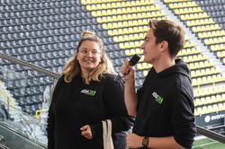 A man holding a microphone and a woman stand in front of a grandstand in a soccer stadium.