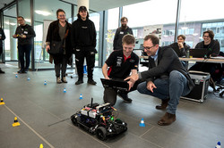 Im Zentrum des Bildes ist ein Auto-Roboter sowie zwei Personen, die sich neben diesen Hinhocken. Im Hintergrund sind weitere Personen zu sehen, die den Blick auf das kleine Roboter-Auto richten.