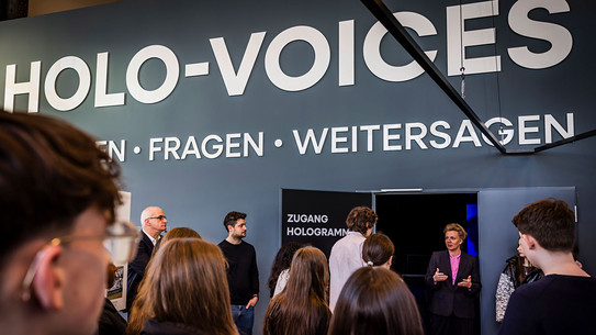 Foto: Mehrere Personen stehen vor einem Eingang mit der Aufschrift „Zugang Hologramme“ unter dem großen Schriftzug „HOLO-VOICES“.