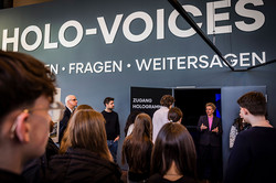 Foto: Mehrere Personen stehen vor einem Eingang mit der Aufschrift „Zugang Hologramme“ unter dem großen Schriftzug „HOLO-VOICES“.