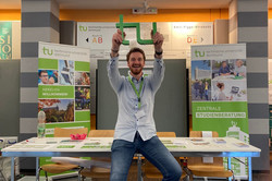 Ein Männlicher Mitarbeiter der Zentralen Studienberatung sitzt auf einem Tisch vor dem Stand der ZSB und hält ein grünes Logo der TU in der Hand.