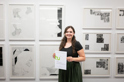 Im Zentrum des Bildes ist Lucy Pawlowicz zu sehen, die mit ihrem Preis-Zertifikat vor einer weißen Wand mit Grafikkunstwerken steht und in die Kamera lächelt.