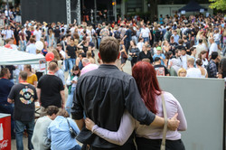 Foto: Beim Sommerfest der TU Dortmund steht ein Paar Arm in Arm und betrachtet das Treiben auf dem Campus. Das Paar hat den Rücken zur Kamera gewendet. Im Hintergrund sieht man die Bühne und Menschen auf dem Sommerfest.
