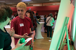 In the photo, you can see two people standing in front of a poster and talking. In the background, other people are standing at high tables.