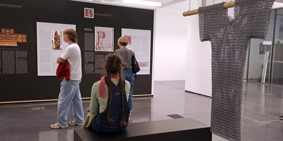 Foto: In einer Ausstellungshalle betrachten mehrere Personen Informationstafeln; eine Person sitzt auf einer Bank, und rechts hängt an einer Stange ein Kettenhemd.