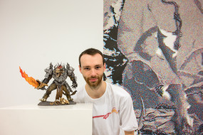Ein Mann hockt hinter einem kleinen Tisch, auf der eine kleine, monsterhafte Figur steht. Hinter dem Mann hängt ein Gemälde an der Wand.