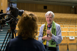 Andreas von Bechtholsheim, ein Mann mit grauen Haaren und hellblauem Hemd, hält ein Mikrofon. Im Vordergrund steht eine junge Frau neben einer Kamera und hält ein Interview mit ihm. Im Hintergrund ein leerer Hörsaal.