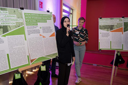 A young woman in a black hijab stands between posters displayed on easels and speaks into a microphone.