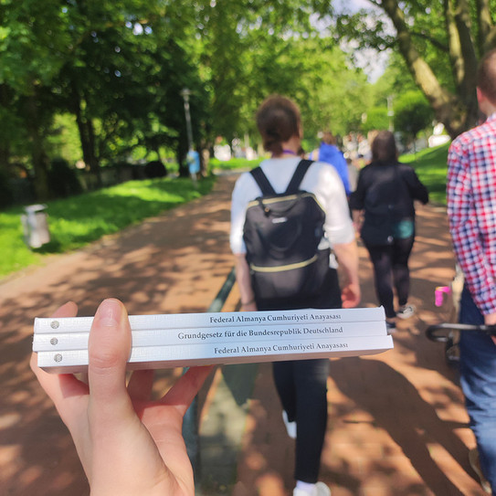 Eine Hand, die drei Bücher in der Hand hält. Im Hintergrund laufen mehrere Personen.
