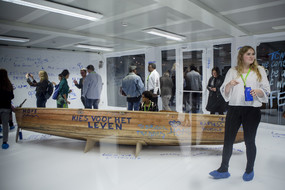 Ein Holzboot steht in einem weiß ausgekleideten Raum mit Menschen. Das Boot und die Wände sind mit blauer Farbe bemalt