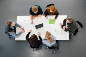  Photographed from above: Six people are sitting around a white table with their laptops