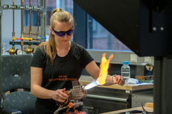 Das Bild zeigt eine schwarz gekleidete Frau mit schwarzer Schutzbrille in einer Werkstatt. Auf ihrem T-Shirt steht "Ich bin Glasapperatebauerin" und hält in einer Hand eine Keule, die sie mit einen Brenner hält.