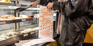 Eine Person nimmt sich aus einer Selbstbedienungstheke ein Stück Kuchen. Sie hält eine Tüte mit der Aufschrift "Gewalt kommt mir nicht in die Tüte". 