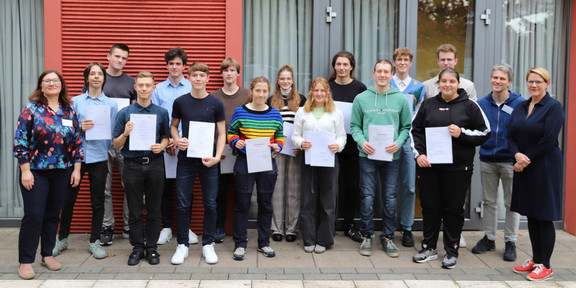 Gruppenbild vor einer roten Gebäudefront: Schüler*innen halten Urkunden in der Hand. Rechts steht Prorektorin Studium Wiebke Möhring mit blonden Haaren und schwarzem Kleid, links steht Dr. Irene Szymanski, eine Frau mit langen braunen Haaren und Brille.