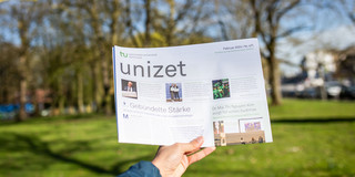 Eine Hand hält die aktuelle Ausgabe der Campuszeitung unizet in die Luft.