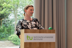 Prof. Tessa Flatten, a woman with blonde hair in a black dress with abstract white flowers, stands at a podium and speaks.