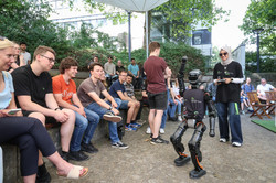Foto: Eine Frau mit Sonnenbrille und Kopftuch steuert einen Robote. Ein neugieriges Publikum schaut dabei zu.
