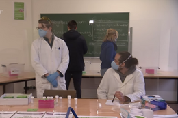 Oktober Eine Gruppe von Menschen steht in einem Raum, zwei haben Arztkittel an.