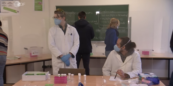 PCR-Tests Eine Gruppe von Menschen steht in einem Raum, zwei haben Arztkittel an.
