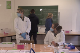 Oktober Eine Gruppe von Menschen steht in einem Raum, zwei haben Arztkittel an.