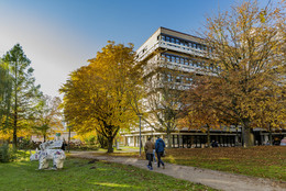 Ein Gebäude der TU Dortmund, Bäume in brauntönen des Herbst stehen vor dem Gebäude. Es laufen zwei Personen vor dem Weg her und links steht eine Nashornfigur auf der Wiese.