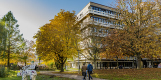 Ein Gebäude der TU Dortmund, Bäume in brauntönen des Herbst stehen vor dem Gebäude. Es laufen zwei Personen vor dem Weg her und links steht eine Nashornfigur auf der Wiese.