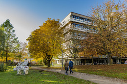 Oktober Ein Gebäude der TU Dortmund, Bäume in brauntönen des Herbst stehen vor dem Gebäude. Es laufen zwei Personen vor dem Weg her und links steht eine Nashornfigur auf der Wiese.