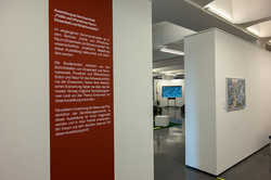Roter Banner mit weißem Text. Der Banner ist auf einer weißen Wand, im Hintergrund sieht man eine Ausstellung.
