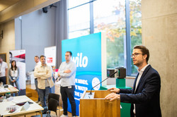 a man is giving a speech at a lectern, people are standing in the background