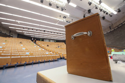 A ballot box stands on a table in the auditorium, people sit on the seats with a large distance between them