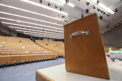 Eine Wahlurne steht auf einem Tisch im Audimax, auf den Plätzen sitzen Menschen mit großem Abstand