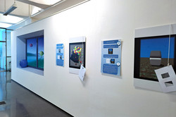 Das Bild zeigt weiße länglich von rechts nach links gehende Ausstellungswand, an der Leinwände hängen. An den Leinwänden hängen jeweils Karten an Schnüren herunter.