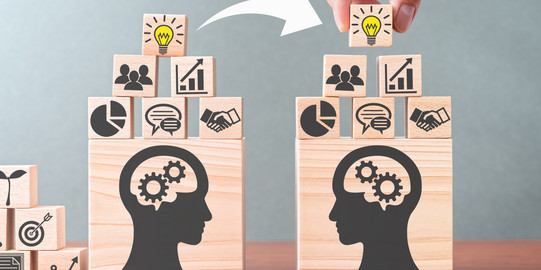 Two towers with identically illustrated blocks stand next to each other. The blocks show icons such as a light bulb, a group of people, a speech bubble, or a pie chart. A white arrow runs from the left to the right tower.