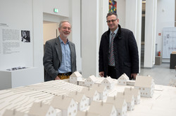 Links ein Mann mit einem grauen Tweed-Jackett und rechts ein Mann mit Brille im schwarzen Anzug.
