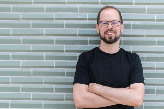 Porträtfoto von Prof. Matthias Feurer: Prof. Feuer trägt ein schwarzes T-Shirt und Brille. Er steht mit verschränkten Armen vor einer grau gemusterten Wand.