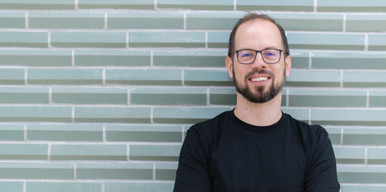 Portrait photo of Prof. Matthias Feurer: Prof. Feurer is wearing a black T-shirt and glasses. He is standing with his arms crossed in front of a gray patterned wall.