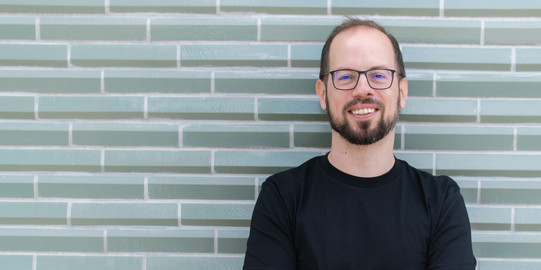Porträtfoto von Prof. Matthias Feurer: Prof. Feuer trägt ein schwarzes T-Shirt und Brille. Er steht mit verschränkten Armen vor einer grau gemusterten Wand.