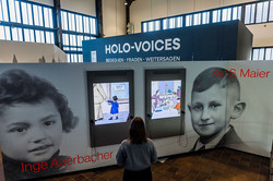 Foto: Eine Person betrachtet interaktive Bildschirme mit Illustrationen in einer Ausstellung mit dem Schriftzug „HOLO-VOICES“.