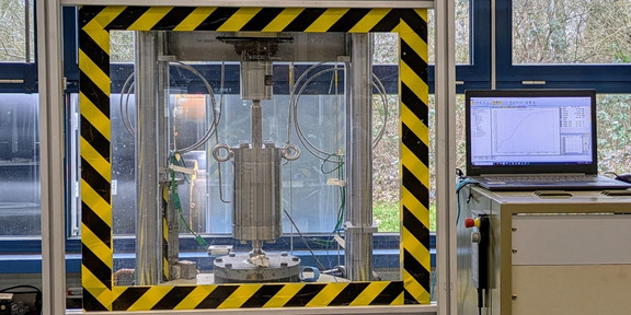 Ein Hochdruck-Wasserstoff-Autoklav hinter einer Glasscheibe, die mit schwarz-gelbem Klebeband umrandet ist. Daneben ein Laptop, der Messungen durchführt.