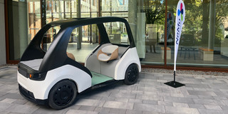 NeMo.bil_Cab Ein offenes Elektro-Gefährt mit zwei gegenüberliegenden Sitzen steht ausgestellt vor einer gläsernen Gebäudefassade. Daneben steht ein Aussteller mit der Aufschrift "NEMO Paderborn".