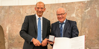 Zwei Herren in Anzug und Krawatte posieren nebeneinander für ein Foto. Der Mann (Prof. Herbert Waldmann) auf der rechten Seite hält eine Medaillen-Schatulle und eine Auszeichnung in den Händen.