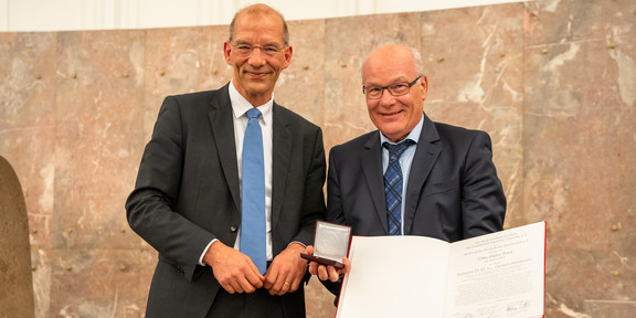 Zwei Herren in Anzug und Krawatte posieren nebeneinander für ein Foto. Der Mann (Prof. Herbert Waldmann) auf der rechten Seite hält eine Medaillen-Schatulle und eine Auszeichnung in den Händen.