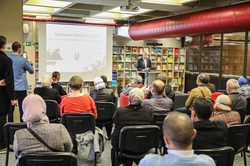 Several people are sitting in the library of the TU Dortmund University listening to a lecture. A man is speaking.