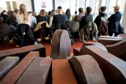 Bricks are placed in the foreground while an audience follows a presentation in the background.