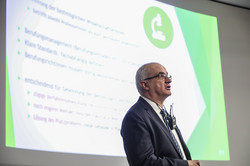 A man gives a lecture, a presentation can be seen in the background