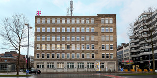 Frontansicht von einem Bürogebäude mit einem Telekom-Logo auf dem Dach. Drumherum sieht man die Straßen und umliegende Gebäude.
