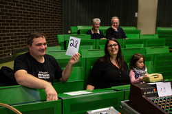 Menschen sitzen in einem Hörsaal. Ein Mann hält eine Nummer in die Höhe.